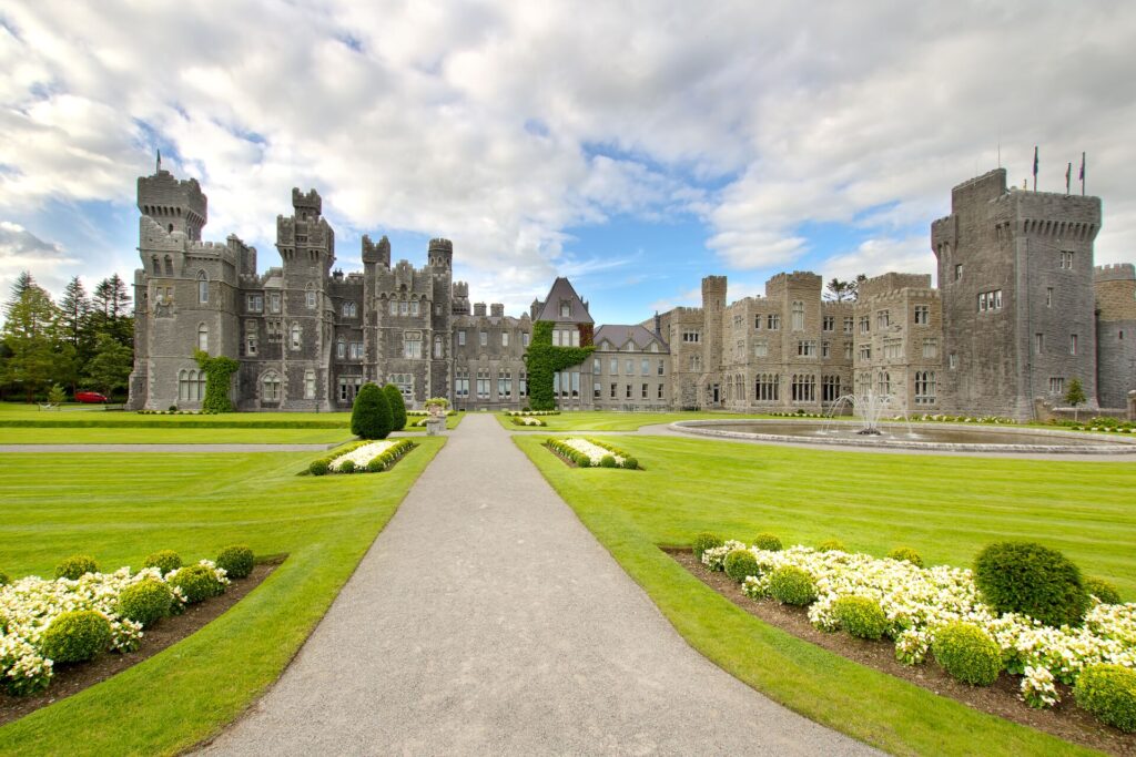 Green lawns and a grey gravel road sweep up to the majestic grey facade and turrets of Ashford Castle, with a bright blue sky filled with fluffy white clouds.