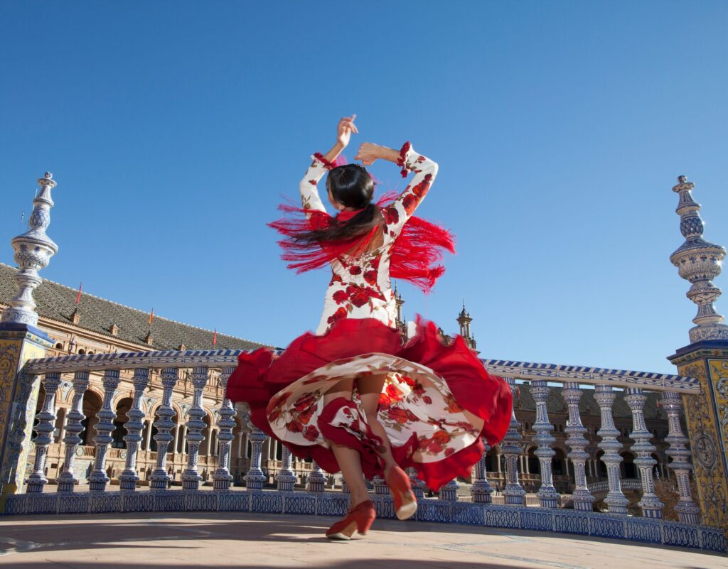 a flamenco dancer in a red and white dress whirls in front of a white and blue tiled bridge barrier, against a bright blue sky.