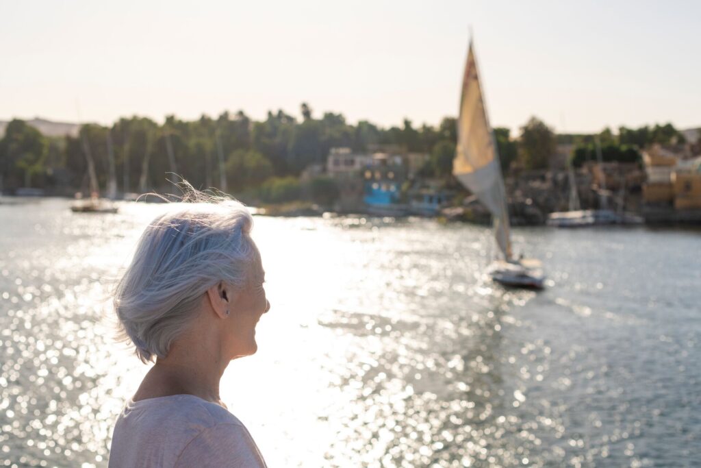 A mature lady looks out over the glistening waters of the River Nile, with a traditional sail boat on the water and blurred buildings on the far banks.