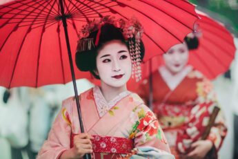 Image of Japanese Geisha wearing kimonos, with traditional make up and red parasols, walking together