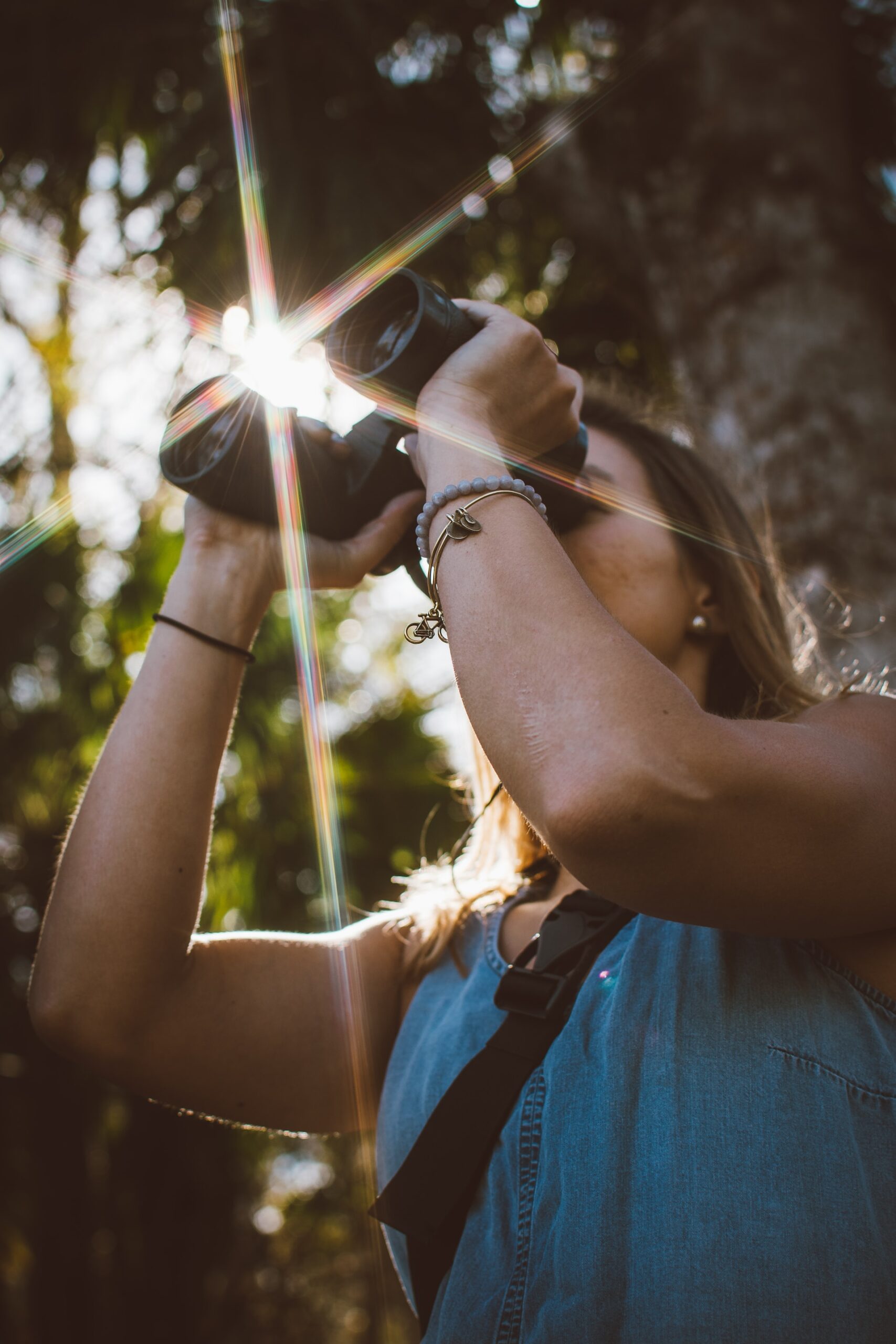 Image of a woman looking through a pair of binoculars, shot from below; sun flaring through the tree canopy above her