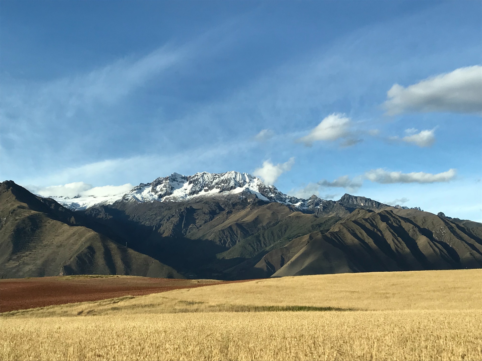 View of the Peruvian Andes, seen from near Cusco. In the foreground, golden rolling field of grass. In the distance, the dark snow-capped body of the Andes rising up through clouds 