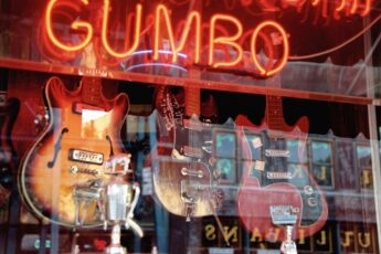 Three guitars are shown in a shop window with a red gumbo sign above them, reflecting the road in a red and orange style picture