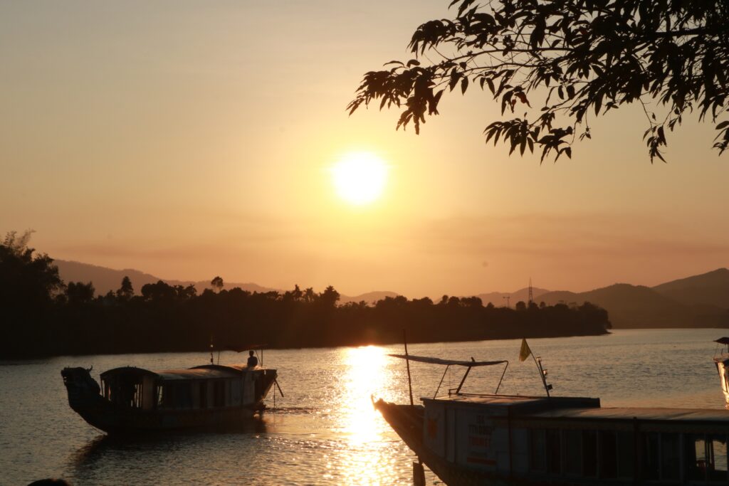 Two wooden boats sail on the rive in Hue, Vietnam with trees in the background and the sun setting.