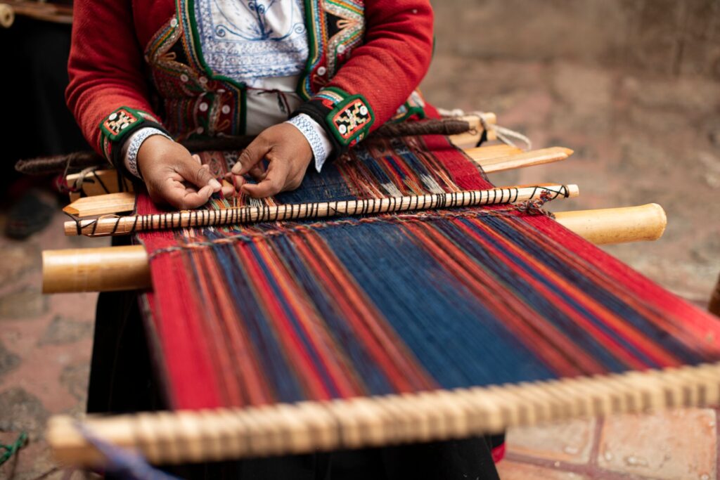 a woman’s hands are shown weaving on a traditional loom in blue and red colours