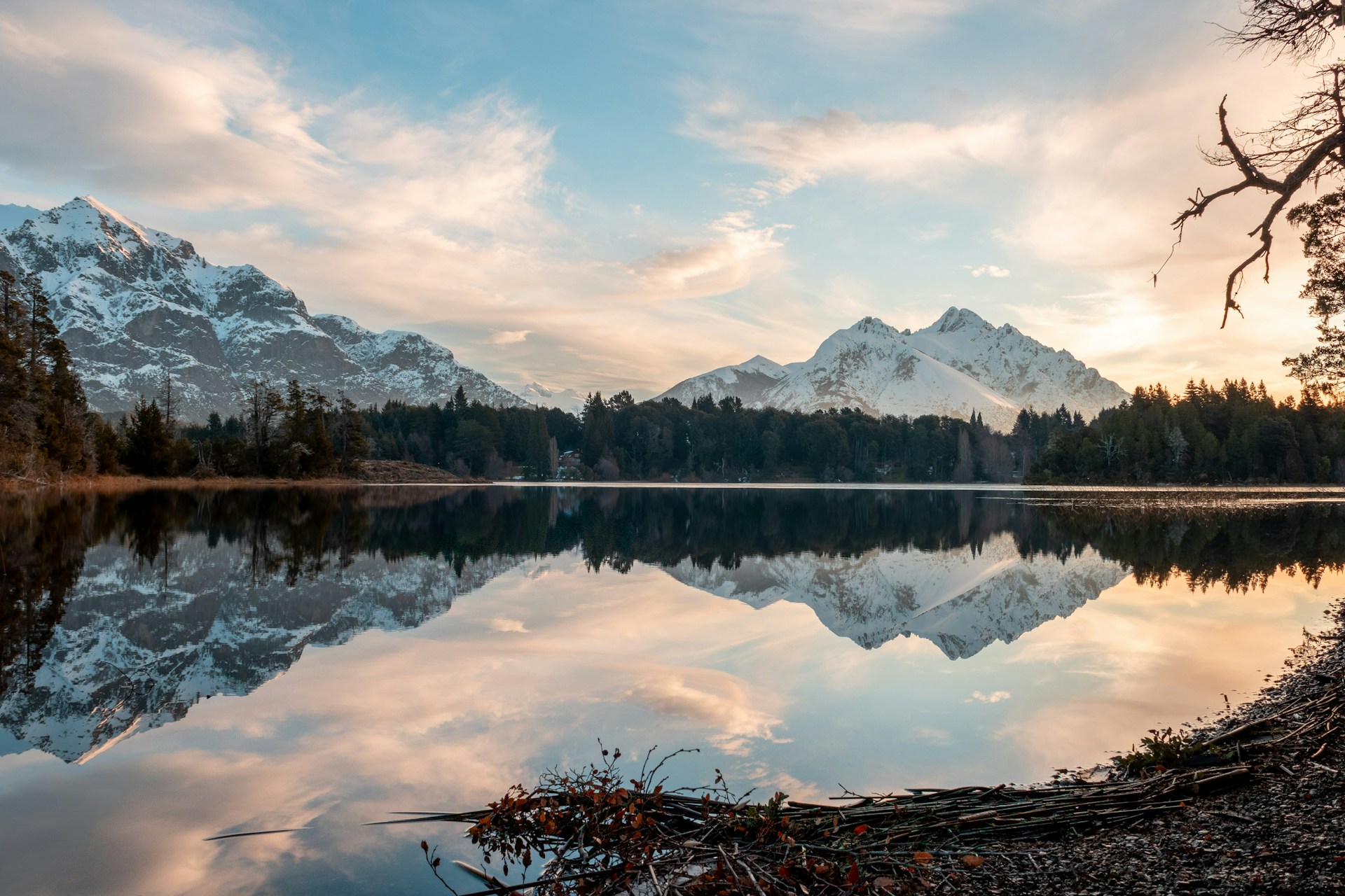 View across a mirror-like lake surface towards jagged, snow-covered mountains. Blue sky with clouds, early morning sun