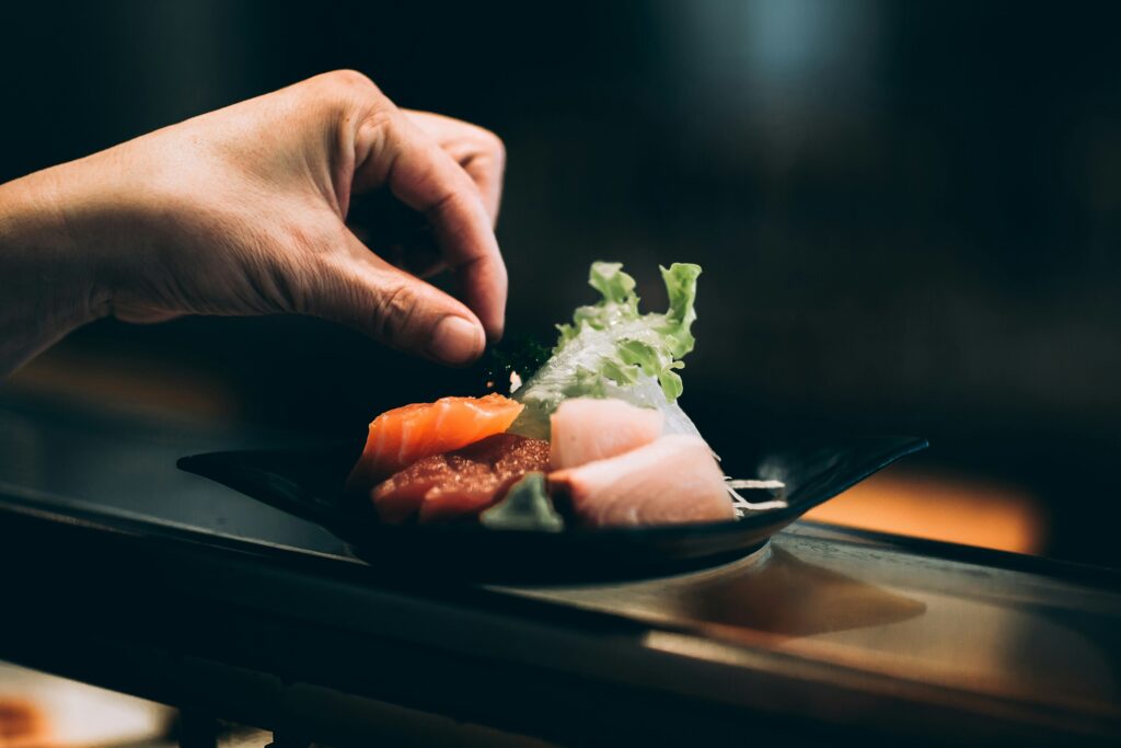 A close up of a hand tending to fine plate of sushi, pinks and greens on a black bowl.