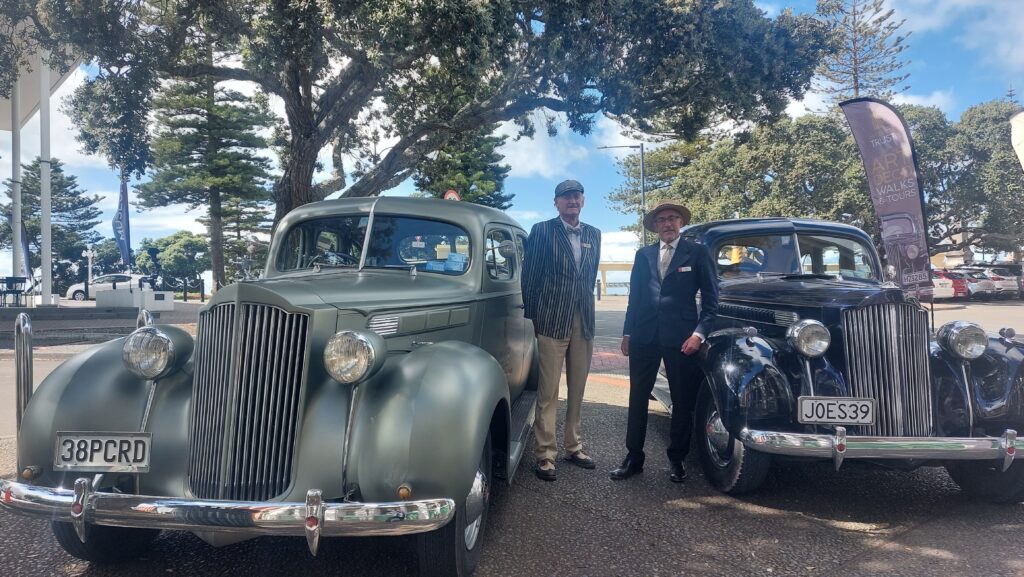 John and colleague stand beside two vintage cars, one green and one dark red under the shade of a tree in Napier, New Zealand
