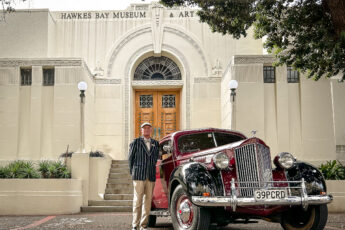 John Ellison stands beside a gleaming dark red vintage car in front of an art deco building in Napier, New Zealand
