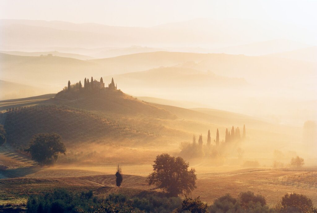 A Tuscan estate sits atop a hill amongst rolling landscape, shrouded in early morning mist and sunlight