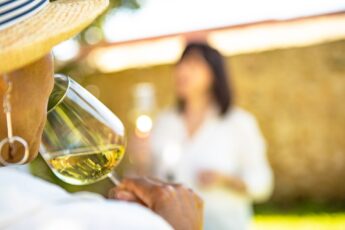 Clos up of lady tasting a white tine, with barred travel partner in the background.