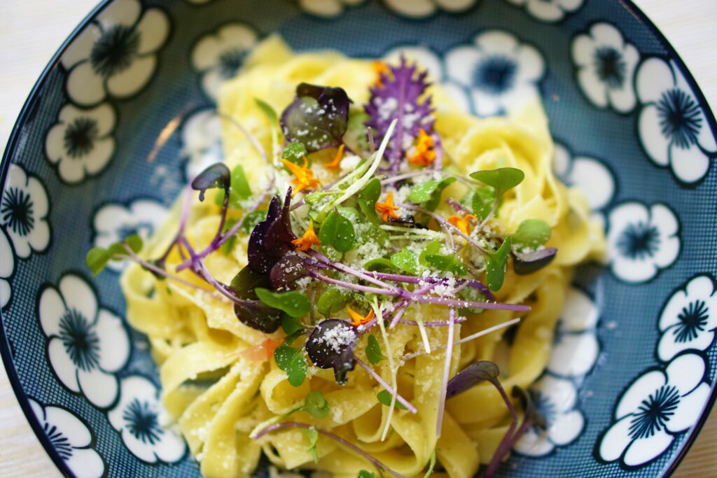 Elegant yellow pasta dish garnished with shaves of dark brown truffle, green and purple salad leaves served in a bowl with a flower pattern.