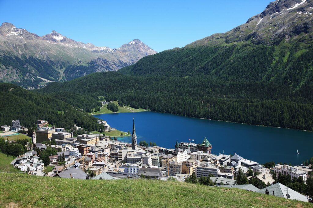 Aerial shot of St Moritz, showing buildings on the edge of a bright blue are with mountains in the background.