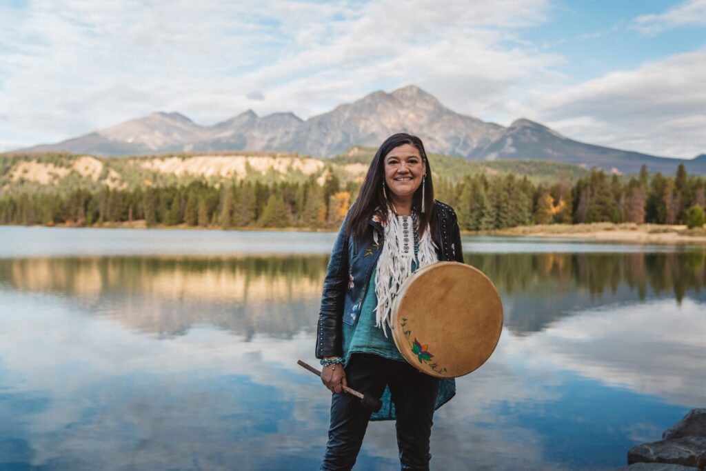 A Warrior Woman stands in front of a large lake with mountains and forrest in the background