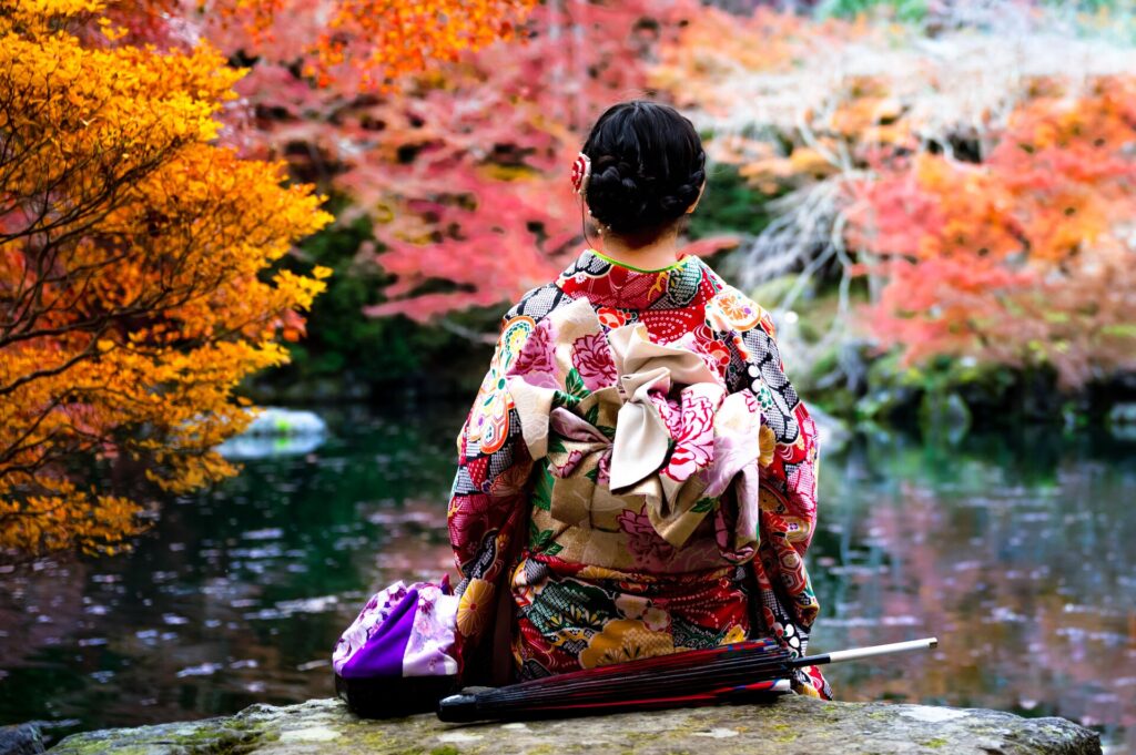 A lady shown from behind in traditional Japanese dress sits in front of a pond with delicate red and pink trees on the far side