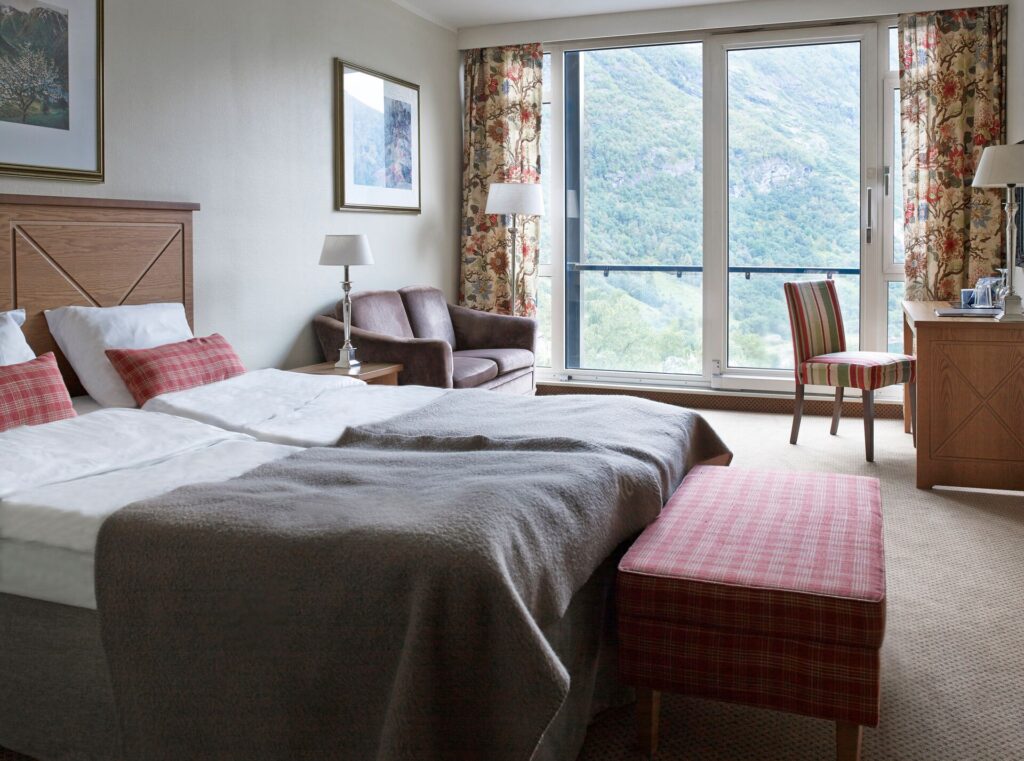 A hotel room is shown with wide windows and a view of rhe Geirangerfjord, with floral curtains and red and white linens.