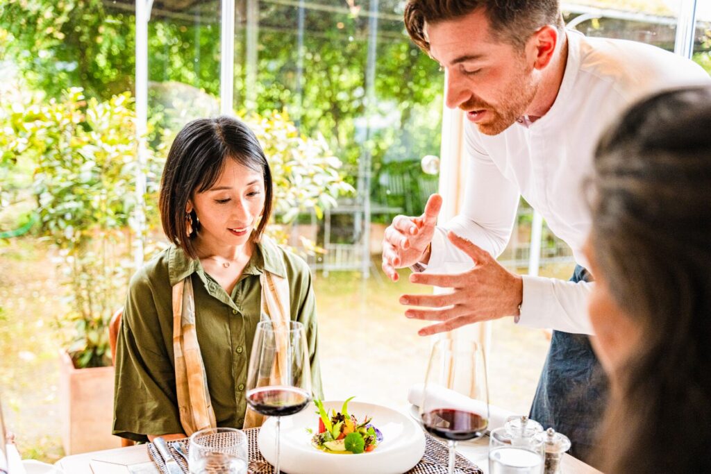 A local chef talks to diners, gesturing over a plate of fine dining.