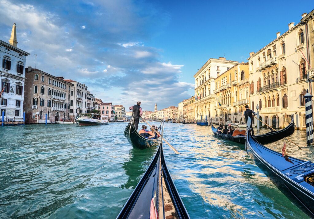 Picture at water level of gondolas on a Venice canal, with green water and golden buildings on the edge and a bright blue sky.