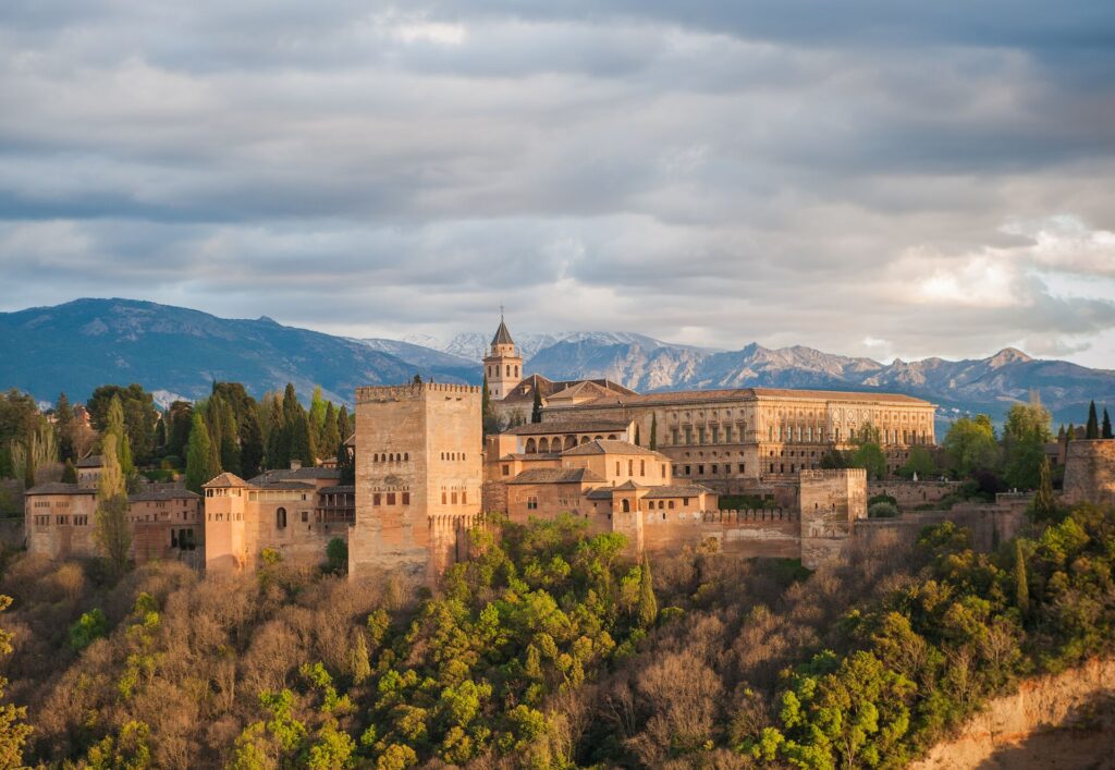 Granada's Alhambra Palace stands proud on a hilltop, beige in colour with grand facades against a cloudy grey sky.