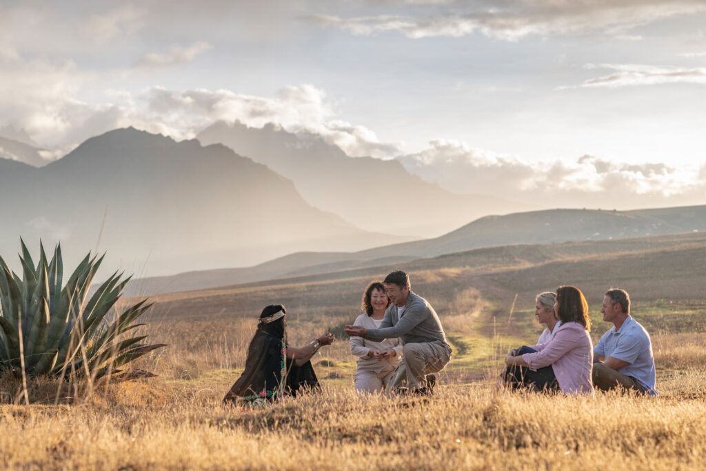 A woman in traditional dress is sat on the ground, with five guests, holding hands out to one of them, with the plains and mountains of Peru's Scared Valley in the background