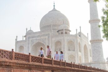 Four guests are pictured, from a distance, walking in front of the huge, white marble Taj Mahal in India, agains a white sky.