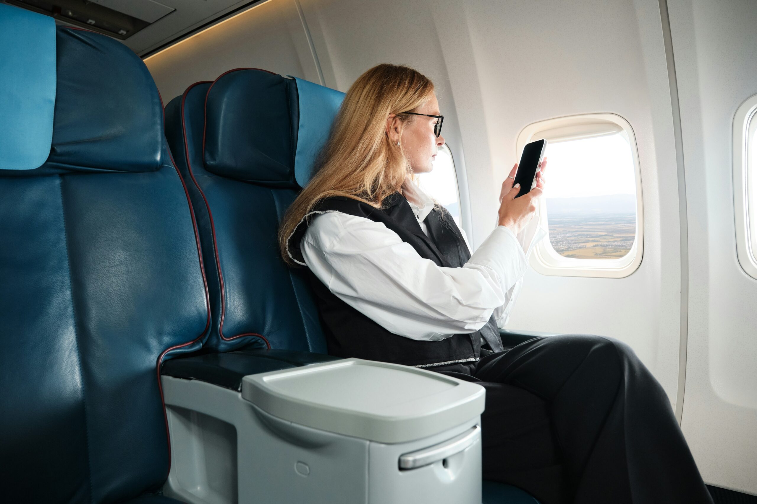 A female passenger sits in an upper class airplane seat, taking a photo on a phone out of the window.