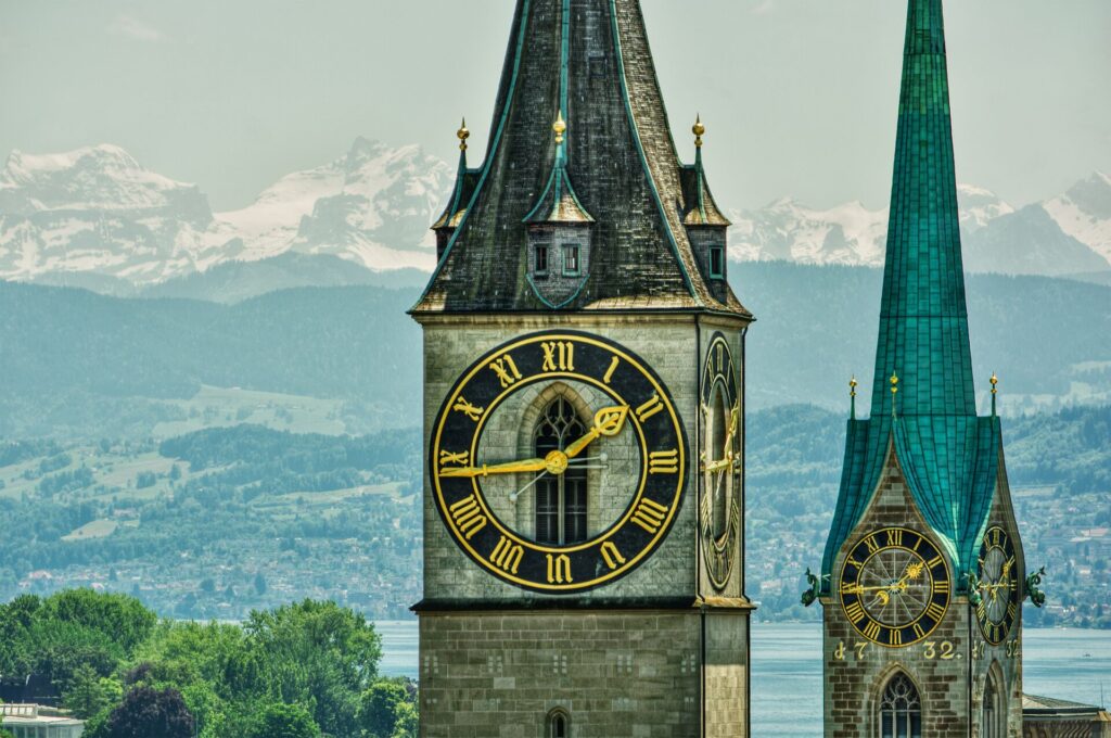 Close up of the clock face atop a tower with snow capped mountains and an emerald green spire behind.