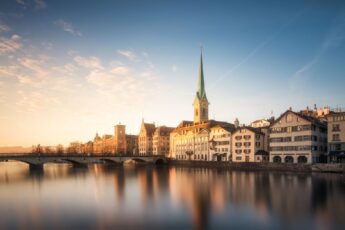 A tall emerald green church spire sits among a row of buildings, with a river an bridge in front of in Zurich, Switzerland.