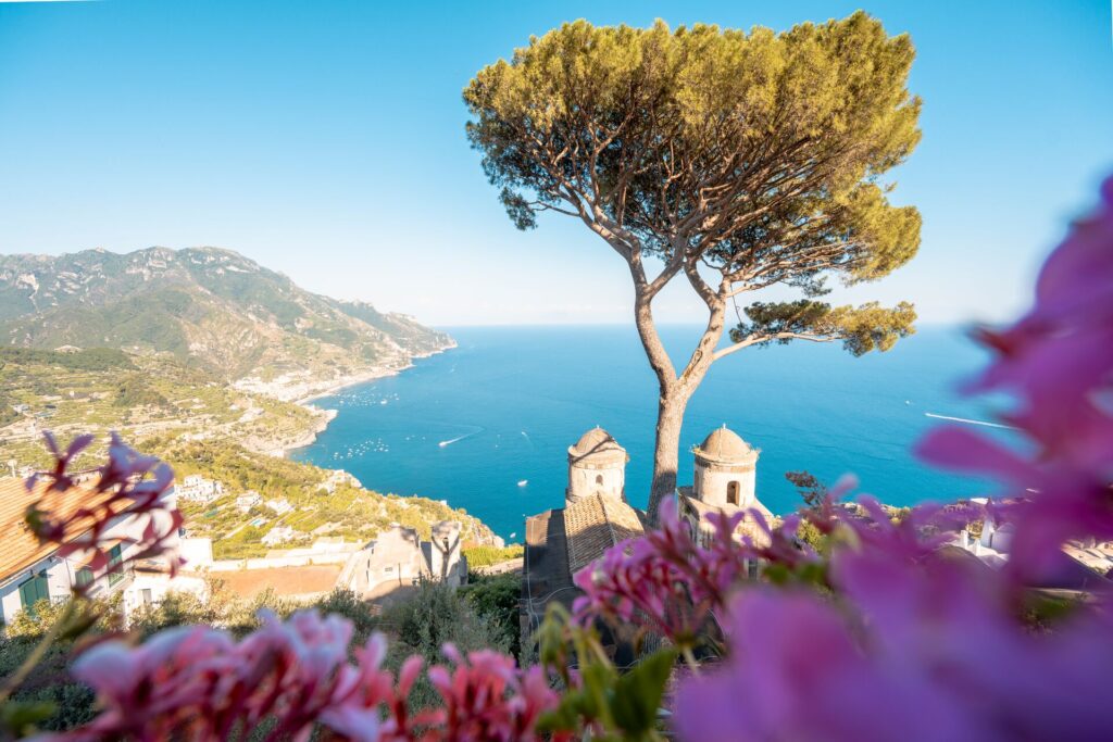 Aerial shot over the cliffs of the Amalfi Coast in Italy, with purple flowers, a bright blue sea and green hillside.
