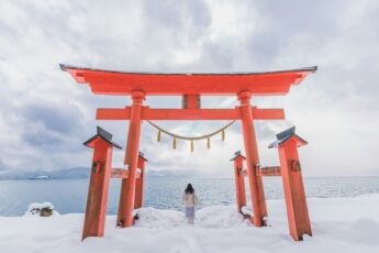 Woman photographed from behind standing beneath torii gate in snow overlooking lake