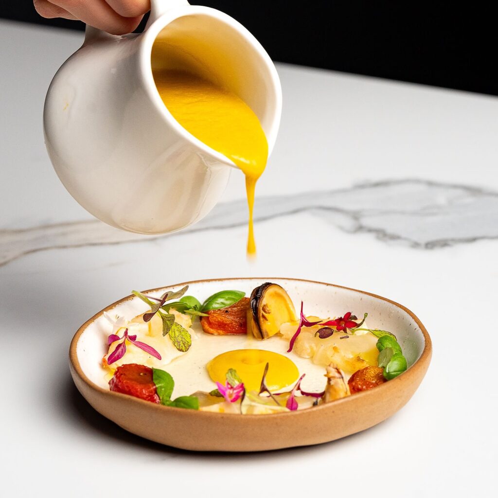 Hand pouring out a jug of yellow sauce onto a colorful plate of food
