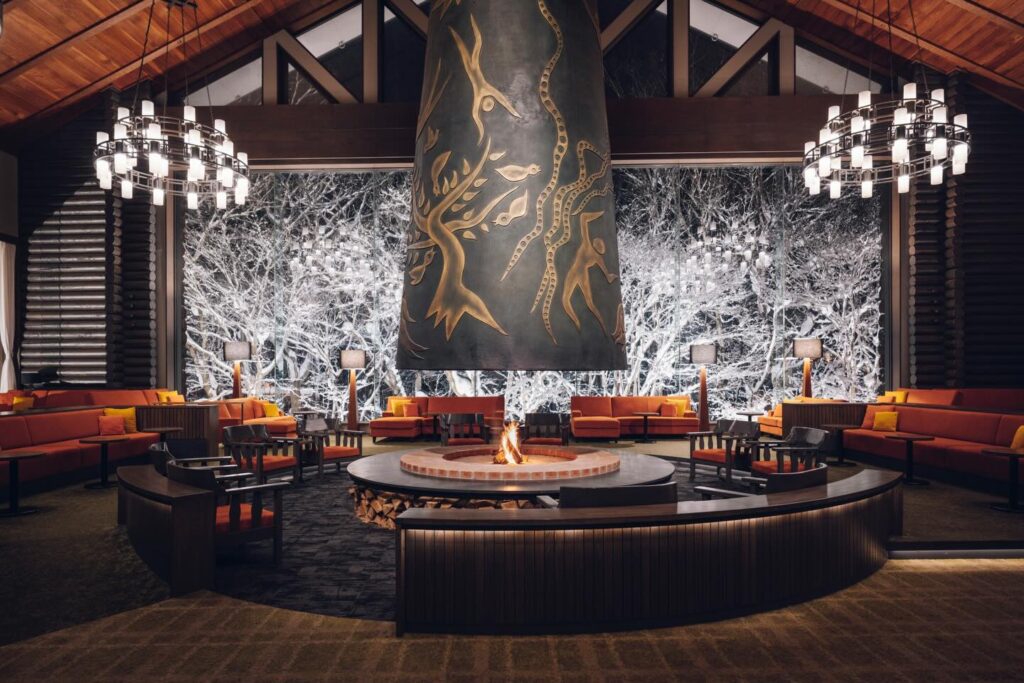 Interior of hotel lounge with circular fireplace in the centre and snow-covered trees beyond the large feature window